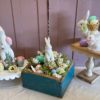 White ceramic bunnies arranged on stands and in a blue wooden box, surrounded by pastel eggs and spring flowers for a cheerful Easter vignette.