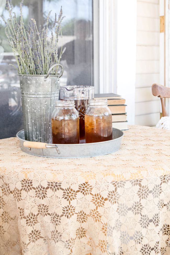 galvanized round metal tray with pitcher of iced tea and two mason jars, accented with a bouquet of lavender in a French flower bucket. Displayed on a lace covered table