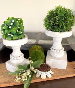 Two white pedestal topiaries with soft green foliage and lily of the valley stems, styled as a simple spring vignette