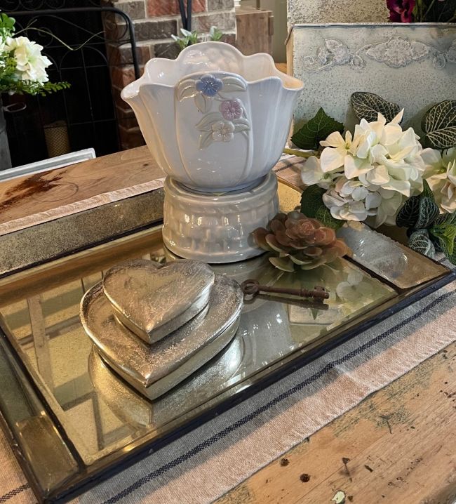 Close‑up of a mirrored tray holding a white embossed floral pitcher, a faux succulent, a vintage key, and two stacked metallic heart boxes, styled on a striped runner with soft spring florals in the background.