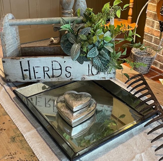 Rustic tabletop vignette featuring a weathered wooden herb box filled with greenery, a mirrored tray with stacked metallic heart boxes, and a vintage metal rake head used as decor, set against a brick and wood background.