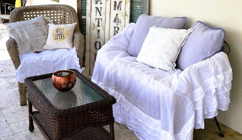 Bright and cozy summer seating corner with a wicker chair, “HELLO SUMMER” pillow, white ruffled settee, and distressed aqua shutters styled outdoors.