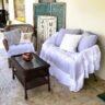 Bright and cozy summer seating corner with a wicker chair, “HELLO SUMMER” pillow, white ruffled settee, and distressed aqua shutters styled outdoors.