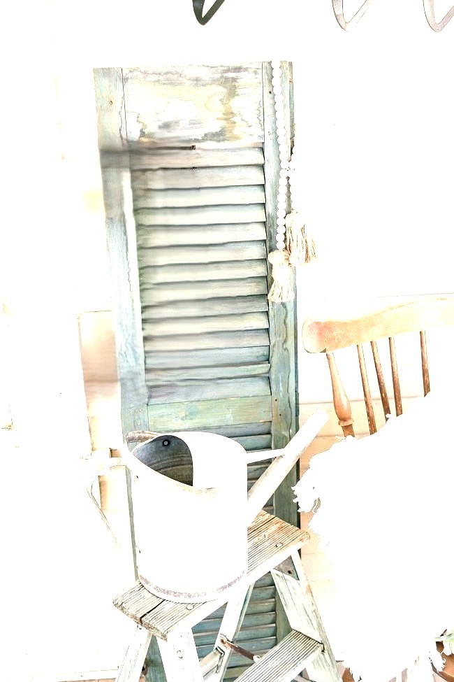 A soft, rustic summer vignette with a weathered shutter, a small wooden stool, a metal watering can, and a light throw draped over a chair, creating a quiet and cozy corner